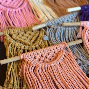 May include: A collection of small macrame wall hangings featuring various colours including pink, cream, tan, mustard yellow, grey, and purple. Each piece is crafted from cotton cord knotted onto a horizontal wooden dowel with a fringe bottom.