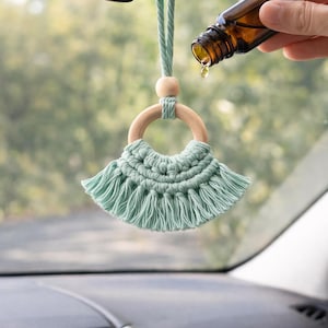 May include: A car air freshener featuring a wooden ring and a mint green macrame design. A brown essential oil bottle is held above the freshener, with a drop of oil falling. The freshener hangs from the rearview mirror inside a car.