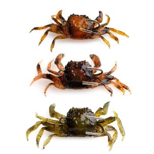May include: Three crab-shaped fishing lures in shades of brown and green. Each lure has a hook attached, designed to mimic a crab. The lures are made of a soft, flexible material.