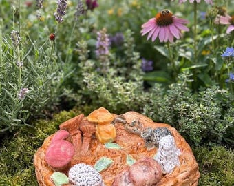 May include: A circular, textured ceramic dish with a light brown, bark-like rim. The interior features various sculpted mushroom shapes in shades of pink, orange, white, and grey, accented with small green leaves scattered across the surface.