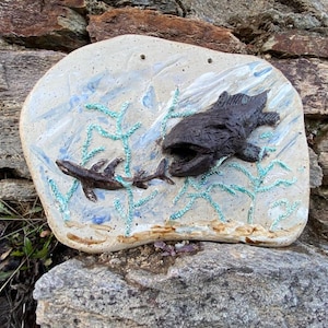 Pode incluir: Um pendente de parede de cerâmica com um peixe castanho texturizado e um peixe menor a nadar através de ondas pintadas de azul e branco. O fundo é de cor castanho claro com algas verdes.