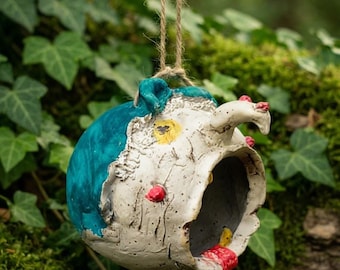 Puede incluir: Una casa para pájaros de cerámica esculpida a mano, de forma redonda, que cuelga de un cordel de yute. El exterior tiene un acabado blanco texturizado con una gran sección azul verdoso, pequeños acentos rojos y una mancha amarilla. Entrada circular.