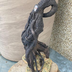 Peut inclure: Une sculpture en argile texturée brun foncé représentant une créature avec une tête tentaculaire et un corps allongé et courbé. La figurine repose sur une base ovale de couleur claire et tachetée, avec des détails de surface poreux.