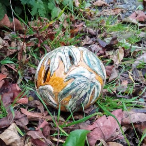 Può includere: Una sfera in ceramica con un design a foglie in tonalità di verde, bianco e arancio. La sfera ha diversi fori tagliati.