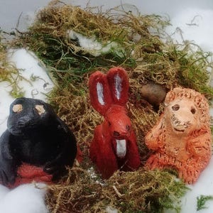 May include: Three small hand-painted clay figurines featuring a black creature with light eyes, a tall red rabbit with white ear accents, and a textured orange creature with a smiling face, all nestled together among green moss and white snow.
