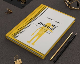May include: A yellow spiral bound journal with the text "My Journal" and "Let's not forget... If I don't write it down I won't remember."  The journal is on a brown leather surface with two pencils and a gold metal decorative stand.