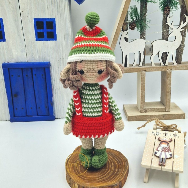 May include: A handmade crochet doll with a festive Christmas theme. The doll wears a green and white striped hat, a green and white striped top, a red skirt, and green boots. The doll is standing on a wooden slice.