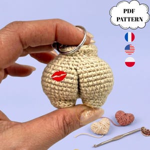 May include: A beige crochet keychain shaped like buttocks with a red embroidered lip print. The item includes a metal keyring and is presented as a PDF pattern. The text PDF PATTERN is visible in a cloud-shaped graphic alongside flag icons.