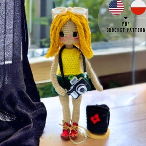 Puede incluir: Patrón de muñeca de crochet en formato PDF. La muñeca lleva un top amarillo, una falda verde y zapatos marrones. Lleva una cámara y una mochila.