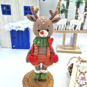 May include: A crocheted reindeer doll with brown antlers, a red nose, and a green scarf. The reindeer wears orange shorts and red mittens, standing on a wooden base. A red heart is attached to the reindeer's chest. A Christmas decoration.