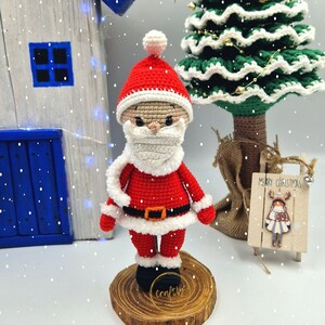 May include: A crocheted Santa Claus figure in a red suit with white trim, a black belt, and a white beard and hat. The figure is standing on a wooden base. The Christmas decoration is handmade. The figure is part of a Christmas display.