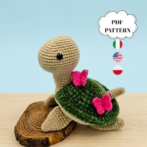 Può includere: Una tartaruga all'uncinetto con testa e arti beige, guscio verde e dettagli a farfalla rosa. La tartaruga è esposta su una piccola base di legno. Una nuvoletta sopra la tartaruga dice "PDF PATTERN".