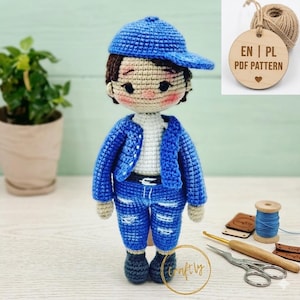 Op de afbeelding: Handgemaakte gehaakte pop van een jongen met een blauwe baseballpet, jas en jeans. De pop heeft bruin haar en een neutrale huidskleur. De pop staat. Een houten label met de tekst "EN | PL PDF PATTERN" hangt op de achtergrond.