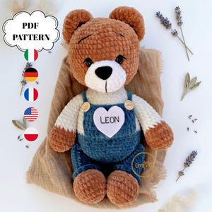 Peut inclure: Un ours en peluche marron fait à la main portant une salopette bleue avec un patch blanc en forme de cœur avec le mot "LEON". L'ours a un nez noir et est assis sur un tissu beige. L'image comprend le texte "PDF PATTERN".
