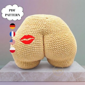 Peut inclure: Un objet en forme de fesses crocheté beige avec une marque de baiser rouge brodée. L'objet a une surface texturée et est présenté sur une surface blanche. Un graphique en forme de nuage avec le texte "PDF PATTERN" est dans le coin supérieur gauche.