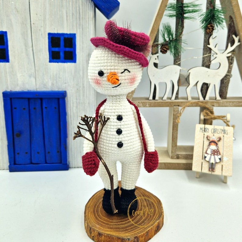 May include: A crocheted snowman figure with a winking expression. The snowman is wearing a burgundy hat, scarf, and mittens, and holding a twig. It stands on a wooden base. The snowman has a carrot nose and black button eyes.
