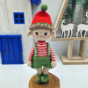 May include: A crocheted elf doll with a red and green hat, a red and white striped top, and green dungarees. The doll has a neutral skin tone and black eyes, and is displayed on a wooden base. A festive Christmas decoration.