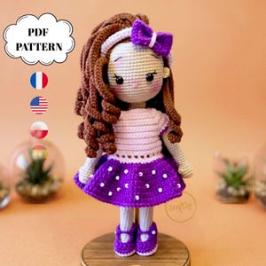 May include: A crochet doll with long brown curly hair, a white headband with a purple bow, a light pink top, and a purple skirt with white polka dots. The doll wears purple shoes and has a light skin tone. Text reads PDF PATTERN.