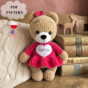 Könnte beinhalten: Ein gehäkelter beigefarbener Teddybär mit einer kleinen rosa Schleife auf dem Kopf, der ein leuchtend rosa Kleid mit einem weißen Herzaufnäher auf der Brust trägt, auf dem der Name MARYSIA steht. Der Text PDF PATTERN ist oben links sichtbar.