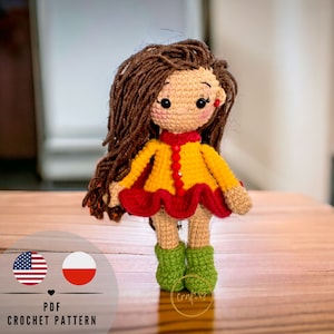 Op de afbeelding: Een handgemaakte gehaakte pop met lang bruin haar, gekleed in een gele jurk met rode rand en groene laarzen. De pop heeft een lachend gezicht met zwarte ogen en rode accenten. De tekst "PDF CROCHET PATTERN" is inbegrepen.
