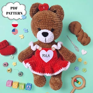 Puede incluir: Un oso de peluche tejido a ganchillo de color marrón que lleva un vestido rojo con un parche de corazón blanco con la inscripción POLA y un cuello de volantes blanco. El oso tiene un lazo rojo en la cabeza y está rodeado de materiales de manualidades.