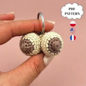 May include: A crochet keychain featuring two cream-colored spheres with brown centers, attached to a silver metal ring. A white cloud-shaped graphic contains the text PDF PATTERN, accompanied by small circular icons of the Polish, American, and French flags.