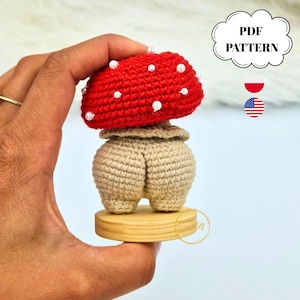 Peut inclure: Une figurine champignon au crochet avec un chapeau rouge vif orné de petites perles blanches et un pied beige en forme de fesses. La figurine est fixée sur une base en bois circulaire. L'image contient le texte PDF PATTERN.