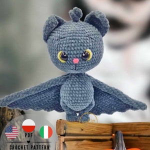 Peut inclure: Un jouet en crochet en forme de chauve-souris amigurumi avec de grands yeux jaunes et un nez rose. La chauve-souris est grise et a de grandes ailes déployées. Le jouet est posé sur une surface en bois.