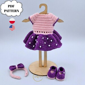 May include: A crochet doll outfit consisting of a light pink bodice with a purple skirt featuring white beads, a matching purple headband with a bow, and a pair of purple shoes. The set is displayed on a wooden hanger and stand. Text reads PDF PATTERN.