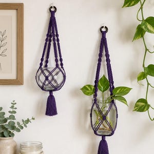 May include: Two purple macrame plant holders, each with a clear glass jar. One jar is empty, the other contains green plants. The holders feature a tassel and a ring for hanging. The design is a decorative way to display plants.