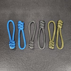 May include: Six cord loops in blue, grey, and olive green. Each loop has a knotted end. The loops are evenly sized and shaped. The background is a dark grey surface. The knotted ends are tightly woven.