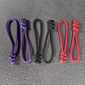 May include: Six zipper pulls made of braided nylon cord arranged in pairs of purple, black, and red. Each pull features a looped design with a knotted end, providing a textured grip for attachment to zips on bags, jackets, or other gear.