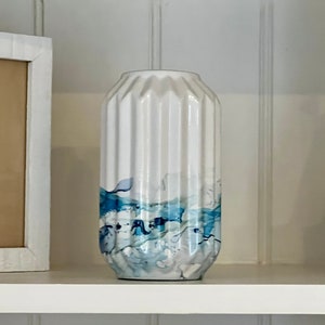 May include: White ceramic vase with vertical ridges and a blue and white abstract design. The vase is cylindrical and sits on a white shelf. A framed seashell art piece and a decorative wooden sailboat are also on the shelf.