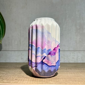 May include: A cylindrical ceramic vase with a vertical fluted texture. The surface features an abstract alcohol ink design in shades of lavender, violet, and deep purple, layered over a white base. The top opening is circular and smooth.