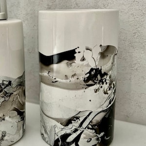 May include: A white ceramic toothbrush holder with black and grey abstract marbling. The holder is cylindrical and has a smooth, glossy finish.