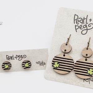May include: Two pairs of wooden earrings with a shamrock design. The larger pair has a striped circle and a smaller plain circle. The smaller pair has a striped circle with a shamrock design. The earrings are displayed on a white card with the text "Pearl+Pego" and "Katie".