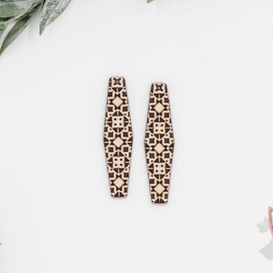 May include: Two wooden earrings with a geometric pattern in brown and cream. The earrings are long and rectangular with rounded ends. The background is white with green leaves.