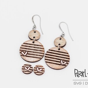 May include: A pair of wooden earrings with a striped pattern and a small heart design. The earrings are made of two circles, one smaller than the other. The earrings are attached to silver hooks. There are also two smaller wooden circles with a heart design, which are likely stud earrings.