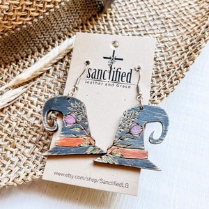 Puede incluir: Un par de pendientes de sombrero de bruja hechos de madera con un diseño de pintura negra y naranja. Los pendientes están decorados con detalles florales y tienen ganchos plateados. Los pendientes se muestran en una tarjeta con el texto "sanctified leather and grace" y una dirección de sitio web.