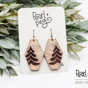 May include: A pair of wooden earrings with a tree design. The earrings are hexagonal in shape and have a natural wood finish. The earrings are on a white card with the text "Pearl + Peg" written in black.