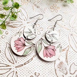 May include: A pair of wooden earrings featuring a floral design. The earrings are painted in shades of pink and green. The earrings are attached to silver hooks.