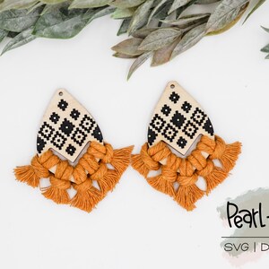 May include: A pair of wooden earrings with a geometric pattern and a tassel fringe. The earrings are painted in black and white, and the tassels are a mustard yellow color. The earrings are displayed on a white background with green leaves.