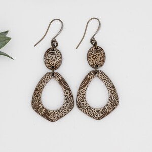 Puede incluir: Un par de pendientes colgantes con un patr&oacute;n floral. Los pendientes est&aacute;n hechos de madera y tienen un acabado marr&oacute;n. Los pendientes est&aacute;n unidos a un gancho plateado.