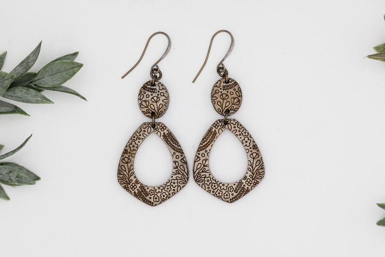 Puede incluir: Un par de pendientes colgantes con un patr&oacute;n floral. Los pendientes est&aacute;n hechos de madera y tienen un acabado marr&oacute;n. Los pendientes est&aacute;n unidos a un gancho plateado.