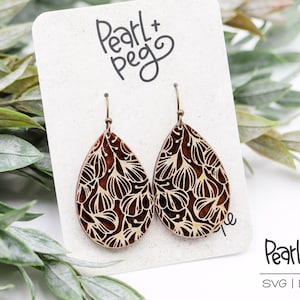 Puede incluir: Un par de pendientes de madera con un diseño floral. Los pendientes son marrones con un patrón floral blanco y tienen ganchos dorados. Los pendientes se muestran en una tarjeta blanca con el texto "Pearl+Peg" escrito en negro.