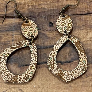 Puede incluir: Un par de pendientes de madera con un patr&oacute;n floral. Los pendientes son marrones con un dise&ntilde;o floral marr&oacute;n claro y tienen forma de l&aacute;grima. Los pendientes est&aacute;n unidos a un gancho dorado.
