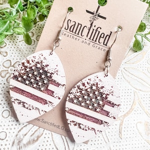 May include: A pair of wooden earrings with a distressed American flag design. The earrings are white with red and blue accents. The earrings are attached to silver hooks. The earrings are from the shop "sanctified leather and grace".