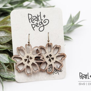 Pode incluir: Um par de brincos de madeira em forma de flores com um design recortado. Os brincos estão em um cartão branco com o texto "Pearl + Peg" escrito nele.