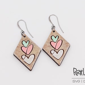 May include: A pair of wooden earrings with a diamond shape and three painted hearts in shades of pink, green, and white. The earrings have silver hooks.