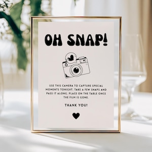 May include: A framed sign with the words "OH SNAP!" in bold, black letters. Below is a black and white illustration of a camera with a heart icon. The sign includes instructions to capture moments and pass the camera. A black heart is at the bottom.
